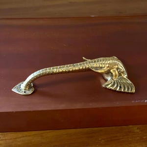 May include: A brass-colored cabinet handle shaped like a fish. The handle curves gently, with a detailed fish head and tail design. The handle is mounted on a dark brown surface.