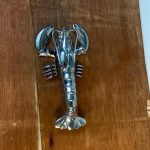 May include: A shiny, silver-colored lobster-shaped bottle opener. The opener is made of metal and has detailed claws, legs, and a segmented body. The lobster is lying on a wooden surface.