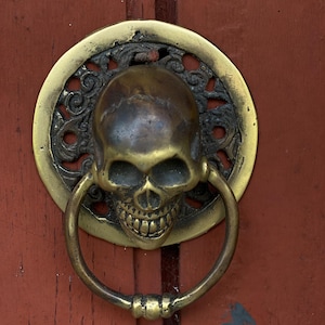 May include: A brass door knocker shaped like a skull with intricate details. The skull has a toothy grin and is surrounded by a decorative ring.