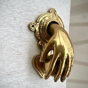 May include: A brass door knocker in the shape of a hand. The hand is holding a round knob and is attached to an ornate backplate. The knocker is mounted on a white wooden door.