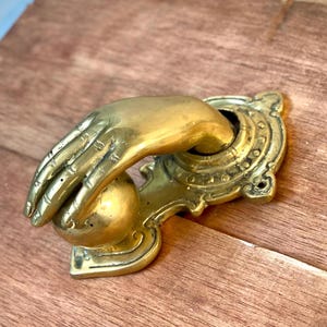 Puede incluir: Un aldaba de latón con forma de mano agarrando una bola. La mano está detallada con nudillos y líneas visibles. La aldaba está montada sobre una placa trasera circular ornamentada con elementos decorativos. El acabado es un oro antiguo cálido.