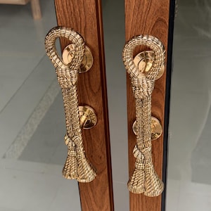 May include: Two vertical door handles made of polished gold-toned metal, each featuring a textured rope design with a looped top and a flared, knotted base. The handles are mounted onto a pair of vertical wooden door frames with a brown wood grain.