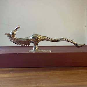 May include: A polished brass figurine shaped like a stylized mythical creature with a long, slender neck, a pointed head, and a curved, textured tail. The object features a central circular detail and stands on a rectangular base.