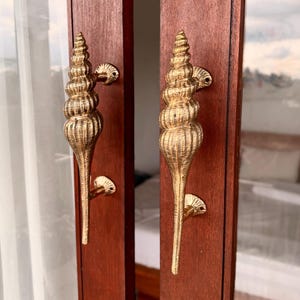 May include: Two gold-coloured door handles, each shaped like a conch shell, are mounted on a dark brown wooden door. The handles have a textured surface and are attached to the door with small, decorative plates.