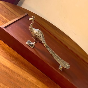 May include: A brass peacock-shaped door handle. The handle is detailed with engraved patterns and features a long tail. The peacock is mounted on a dark brown wooden surface, with a warm, inviting colour palette.