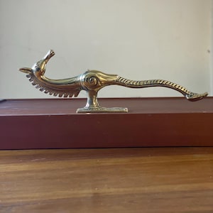 May include: A brass sculpture of a stylized bird with a long, curved tail. The bird has a detailed head and a textured body. The sculpture is mounted on a small base and is displayed on a wooden surface.