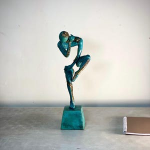 Könnte beinhalten: Eine abstrakte Skulptur in Türkis und Gold, die eine Figur in einer kontemplativen Pose darstellt. Die Skulptur ist auf einem quadratischen Sockel montiert und hat eine strukturierte Oberfläche. Die Form der Figur ist stilisiert, mit länglichen Gliedmaßen und einem runden Kopf. Die Skulptur steht auf einer grauen Oberfläche.
