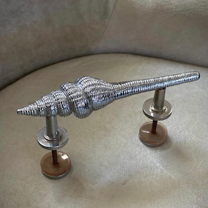 May include: A polished silver seashell-shaped door handle. The handle is mounted on two posts with circular bases. The seashell design features detailed ridges and a pointed end. The handle is a decorative element for doors or furniture.