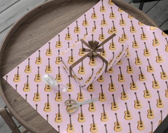 May include: A wrapped gift box and wrapping paper featuring a repeating pattern of acoustic guitars on a pink background. The gift is tied with a brown ribbon. Scissors and tape are also on the table.