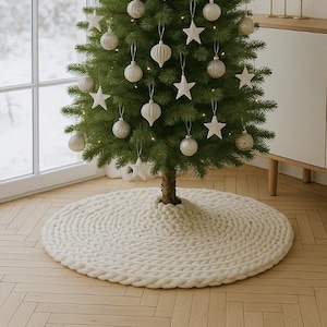 Op de afbeelding: Een versierde kerstboom met witte ornamenten en stervormige decoraties. De boom staat op een crèmekleurige, cirkelvormige gebreide boomrok. De boom staat op een lichtgekleurde houten vloer.
