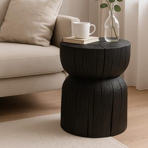 May include: A black, wooden side table with a unique, stacked design. A white mug and a book sit atop the table. A clear glass vase with greenery is also on the table. The table is next to a beige sofa.
