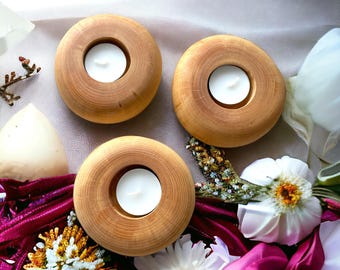 Può includere: Tre portacandele in legno dal design circolare, ciascuno con una candela bianca a lumino. I portacandele sono in legno chiaro e sono disposti su una superficie con fiori e tessuto.