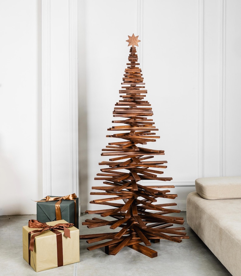 May include: A tall, conical Christmas tree sculpture constructed from stacked, horizontal dark brown wooden slats. The structure features a central vertical support and is topped with a small, eight-pointed wooden star ornament.