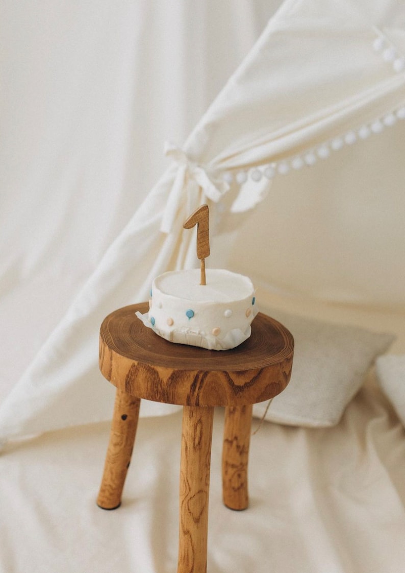 Op de afbeelding: Een kleine witte cake met blauwe en witte sprinkles en een houten nummer &eacute;&eacute;n erop, die op een houten kruk staat.