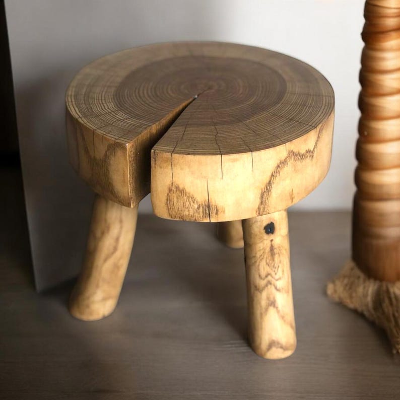 Op de afbeelding: Een houten kruk met een rond, natuurlijk houten blad en drie poten. Het blad is gemaakt van een boomstamschijf, wat de natuurlijke nerf en ringen laat zien.