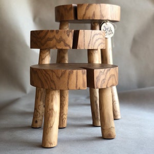 Op de afbeelding: Twee houten krukjes met een natuurlijke afwerking. De krukjes zijn op elkaar gestapeld. Het bovenste krukje heeft een klein label met de tekst "Spiz & Jana".