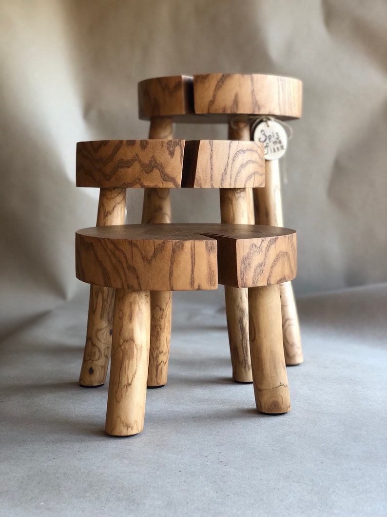 Op de afbeelding: Twee houten krukjes met een natuurlijke afwerking. De krukjes zijn op elkaar gestapeld. Het bovenste krukje heeft een klein label met de tekst "Spiz & Jana".