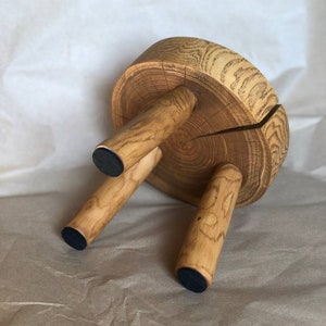 Op de afbeelding: Een houten kruk met een ronde bovenkant en drie poten. De bovenkant van de kruk is gemaakt van een boomstamschijf, en de poten zijn gemaakt van kleinere stukken hout. De kruk heeft zwarte rubberen voetjes aan de onderkant van elk pootje.