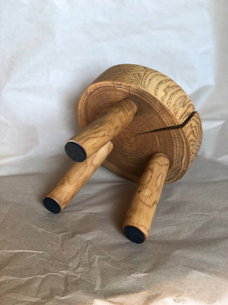 Op de afbeelding: Een houten kruk met een ronde bovenkant en drie poten. De bovenkant van de kruk is gemaakt van een boomstamschijf, en de poten zijn gemaakt van kleinere stukken hout. De kruk heeft zwarte rubberen voetjes aan de onderkant van elk pootje.