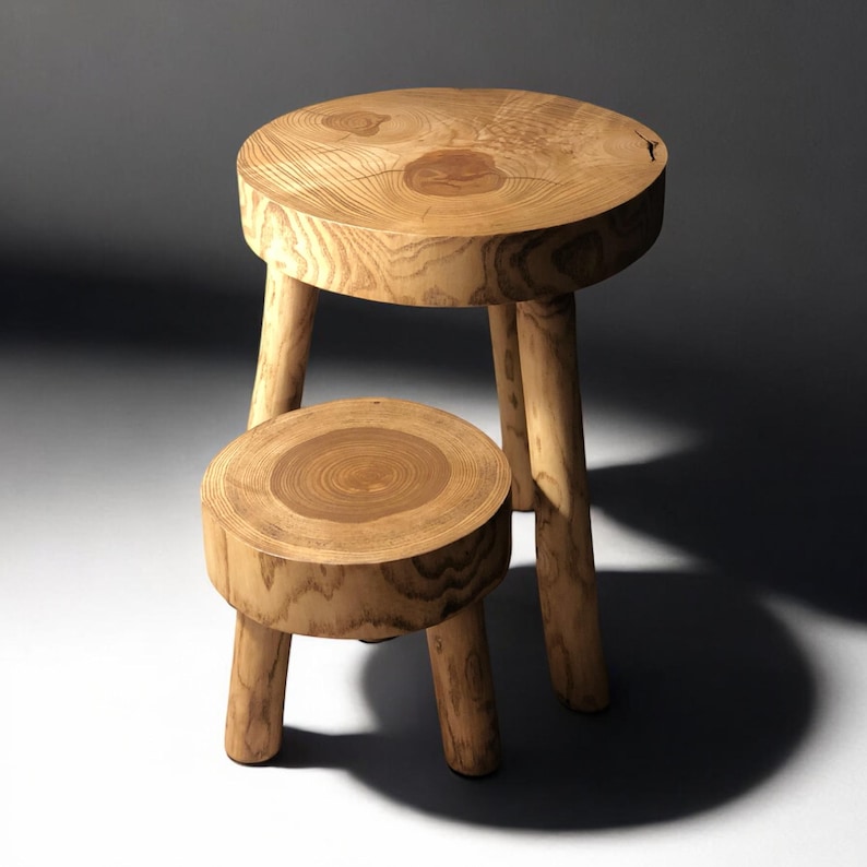 Op de afbeelding: Twee houten krukjes, een groter dan de andere, met ronde bovenkanten en drie poten. De krukjes zijn gemaakt van natuurlijk hout met een lichtbruine kleur en een nerfpatroon.