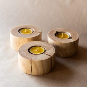 May include: Three wooden candle holders with a natural wood finish. Each holder has a circular opening to hold a small tea light candle. The candles are lit and glowing.