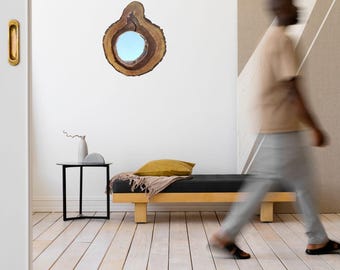 May include: A wooden mirror with a natural, organic shape hangs on a white wall. The mirror's frame is a cross-section of a tree trunk, showcasing the wood's grain. A person walks past a daybed with a yellow pillow.