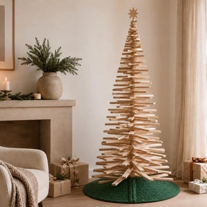 May include: A tall, conical Christmas tree constructed from stacked horizontal light-colored wooden slats, featuring a star-shaped topper and resting on a circular, dark green crocheted tree skirt on the floor.