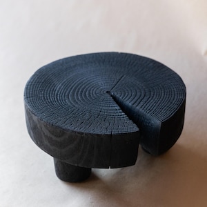 May include: A black wooden pedestal with a round top and three legs. The top is made of a single piece of wood with a natural grain pattern.