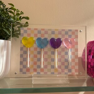 May include: A clear acrylic frame containing a rectangular print with a pastel checkered pattern and four heart-shaped lollipops in yellow, blue, purple, and pink. The frame is supported by a clear acrylic stand on a glass surface.