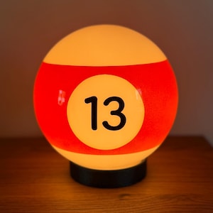 May include: A spherical lamp shaped like a number 13 billiard ball. The lamp features a white base color with a wide horizontal red stripe encircling the middle. A white circular patch in the center displays the black number 13. The lamp glows.