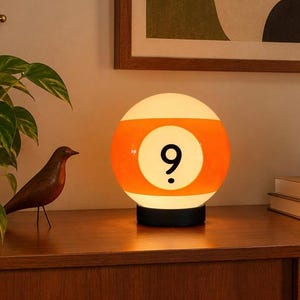 May include: A spherical table lamp designed to resemble a yellow and white billiard ball with the number nine printed in black on a white circular background. The lamp features a smooth, glowing surface and rests on a circular black base.