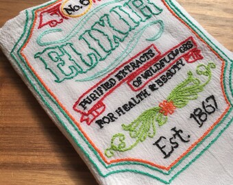 May include: White kitchen towel with embroidered text that reads "No. 6 Elixir Purified Extracts of Wildflowers For Health & Beauty Est. 1867". The embroidery is in green, orange, red, and black thread.