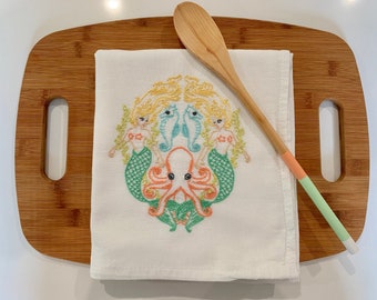 May include: A white kitchen towel with an embroidered design of two mermaids, two seahorses, and an octopus. The design is in shades of green, yellow, orange, and pink.