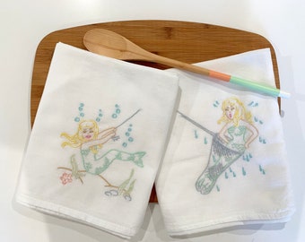 May include: Two white kitchen towels with embroidered images of mermaids. One mermaid is holding a fishing rod and the other is holding a net. Both mermaids have long blonde hair and are wearing green tails.