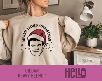 Könnte beinhalten: Ein beigefarbenes Sweatshirt mit Rundhalsausschnitt und einer schwarzen Strichzeichnung eines Mannes mit roter Weihnachtsmütze. Der Text "MERRY LLOYD CHRISTMAS" ist in einem Bogen darüber gedruckt, ergänzt durch kleine Sterne und Schneeflocken.