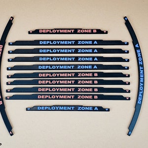 Könnte beinhalten: Ein Satz von 12 schwarzen und weißen Spielsteinen mit dem Text "Deployment Zone A" und "Deployment Zone B", der in Rosa und Blau gedruckt ist. Die Steine sind gebogen und haben ein spitzes Ende.