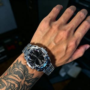 May include: A silver-toned wristwatch featuring a metallic band and a circular case with a black dial. The bezel and lugs are designed with intricate, sharp, claw-like protrusions that extend over the edges of the watch face, creating a bold, textured appearance.