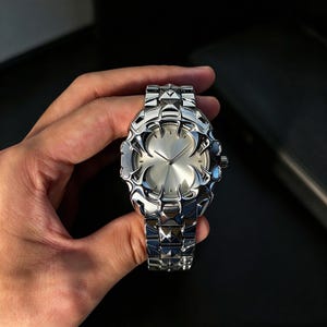May include: A silver-toned wristwatch featuring a metallic band with pyramid-shaped links. The watch face has a smooth, circular centre surrounded by a decorative, claw-like frame with sharp, pointed edges and a polished, reflective finish.