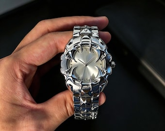 May include: A silver-toned wristwatch featuring a metallic band with pyramid-shaped links. The watch face has a smooth, circular center surrounded by a decorative, claw-like frame with sharp, pointed edges and a polished, reflective finish.