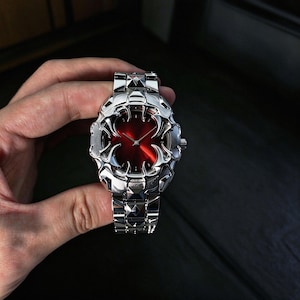 Peut inclure: Une montre-bracelet argentée dotée d'un cadran circulaire rouge métallique. Le boîtier et le bracelet présentent une conception complexe, superposée et angulaire. Les maillons du bracelet ont une texture facettée en forme de pyramide.