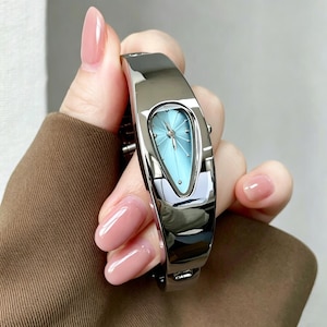 Puede incluir: Un reloj tipo brazalete plateado con una banda de metal pulido y reflectante y una esfera en forma de lágrima. La cara del reloj es de un color azul claro suave con manecillas plateadas y un pequeño marcador en la punta inferior de la lágrima.