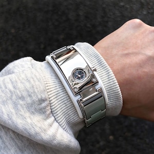 Könnte beinhalten: Eine Armbanduhr mit einer breiten, polierten, rechteckigen Metallplatte in Silberoptik und einem kleinen, runden analogen Zifferblatt in der Mitte. Die Uhr hat einen weißen Rahmen und ein segmentiertes Metallarmband mit seitlicher Krone.