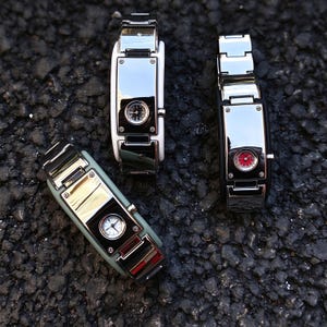 May include: Three rectangular wristwatches with metallic silver-tone faces and polished frames. One watch features a white dial, one a red dial, and one a black dial. Each watch is set into a distinct colored band: white, black, and sage green.