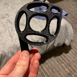 Peut inclure: Un composant circulaire noir imprimé en 3D présentant une surface texturée et plusieurs découpes ovales de tailles variées. L'objet comprend une petite languette saillante sur le dessus et un bras plat et droit s'étendant du bord inférieur.