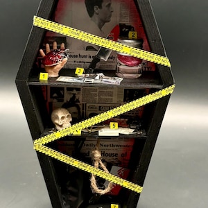 Puede incluir: Una vitrina negra en forma de ataúd con múltiples estantes, que presenta elementos de escena del crimen en miniatura. Incluye una mano sosteniendo un corazón, un cuchillo, un frasco con un cerebro, una calavera, una soga y cinta de escena del crimen.