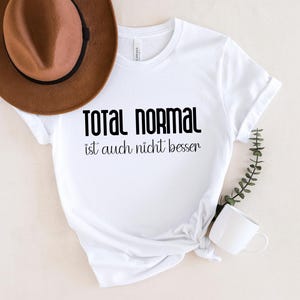 May include: White t-shirt with black text that reads "Total Normal ist auch nicht besser".
