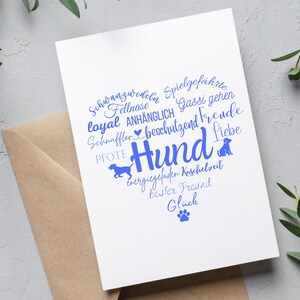 May include: A white greeting card featuring a heart-shaped word cloud in blue ink. The text includes German words like Hund, Liebe, and Pfote, alongside silhouettes of dogs and a paw print. The card is placed on a brown envelope.