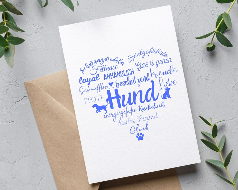 May include: A white greeting card featuring a heart-shaped word cloud in blue ink. The text includes German words like Hund, Liebe, and Pfote, alongside silhouettes of dogs and a paw print. The card is placed on a brown envelope.