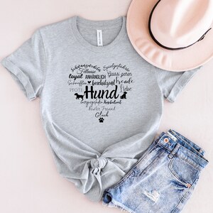 May include: A heather gray short-sleeve t-shirt featuring a black graphic print of a heart shape formed by German words related to dogs, including the word Hund, along with small dog silhouettes and a paw print icon, paired with a pink hat and denim shorts.