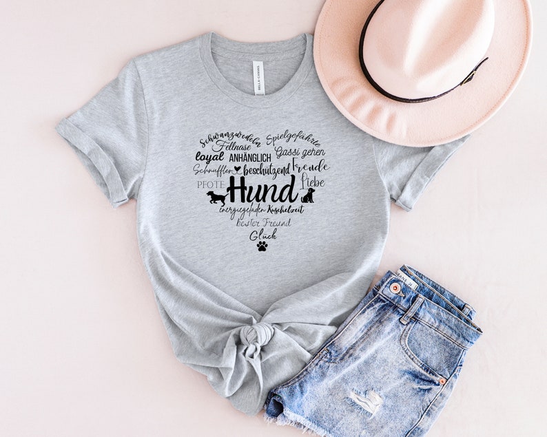 May include: A heather gray short-sleeve t-shirt featuring a black graphic print of a heart shape formed by German words related to dogs, including the word Hund, along with small dog silhouettes and a paw print icon, paired with a pink hat and denim shorts.